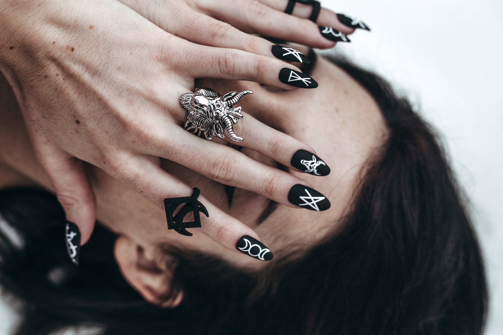 girl with black hair with hands over face wearing goat head ring and satanic matte black stiletto fake nails