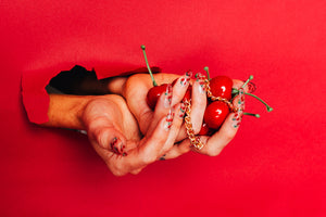 hand exploding through red paper holding cherries and wearing press on nails with a cherry print and a gold chain attached