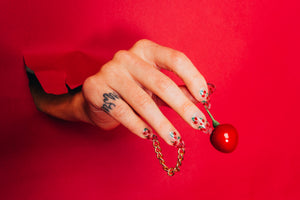 a hand with a flame tattoo bursting through red background holding a cherry wearing clear cute press on nails