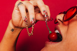 girl eating a cherry wearing clear press on nails that are attached by a chain and have cherries on them