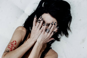 girl with black hair and tattoos with hands covering face wearing matte black almond shaped fake nails with witchy symbols